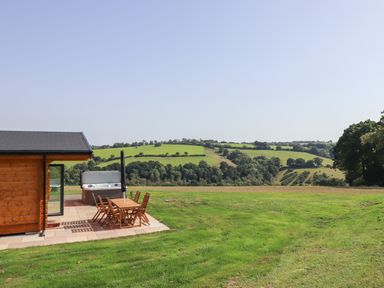 A wooden cabin patio with table chairs and a hot tub overlooking green fields at Foxglove Lodge in Hittisleigh