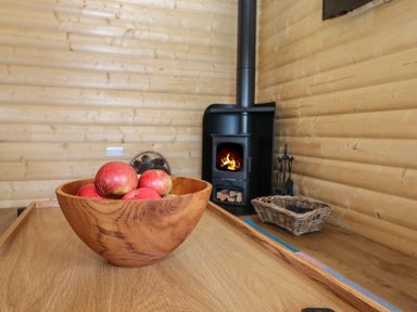 A wooden bowl with apples on a table near a black stove and a wicker basket in a wooden room at Bluebell Lodge in Hittisleigh