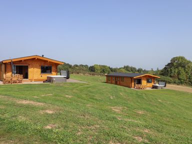 Two wooden lodges with outdoor hot tubs and patio furniture on a grassy hill with trees in the background at Bluebell Lodge in Hittisleigh