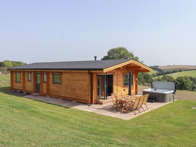 A wooden lodge with outdoor dining table and hot tub on a patio in a grassy rural setting at Bluebell Lodge in Hittisleigh