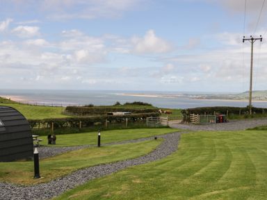 A grassy garden with a gravel path and a wooden picnic table overlooking a coastal landscape with water and hills at Orchard in Dalton-In-Furness