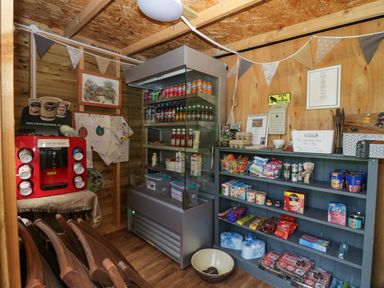 A small wooden shop interior with a refrigerator, shelves stocked with snacks and drinks, a hot water dispenser, and folded wooden chairs at Orchard in Dalton-In-Furness