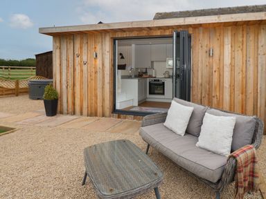An outdoor seating area with a wicker sofa and table in front of a wooden cabin with open doors showing a kitchen at Hillside Hut Radbourne near Ashbourne