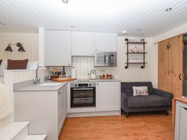 A compact kitchen with white cabinets a built-in oven microwave and sink next to a dark gray loveseat with a floral cushion at Hillside Hut in Radbourne near Ashbourne