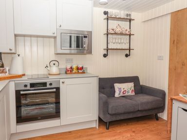 A kitchen area with white cabinets oven microwave kettle and a dark gray sofa with a floral cushion at Hillside Hut Radbourne near Ashbourne