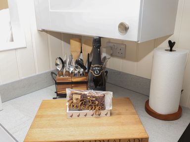 A kitchen countertop with utensils in a wooden holder a bottle a paper towel roll and a wooden cutting board with a welcome sign and packaged food at Hillside Hut in Radbourne near Ashbourne