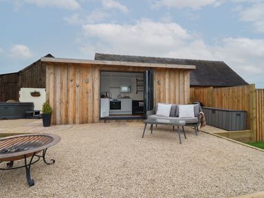 A wooden cabin with open doors showing a kitchen and seating area outside with a sofa table and fire pit at Hillside Hut in Radbourne near Ashbourne