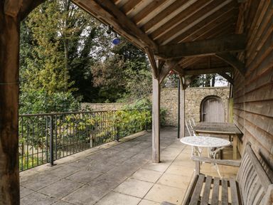 An outdoor covered patio with wooden bench wooden table white metal chairs and a stone wall with an arched wooden door at The Barn at Westhall Cottage Fulbrook near Burford