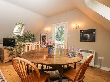 A dining area with a round wooden table and chairs a vase with flowers on the table a TV on a wooden stand plants and wall lights at The Barn at Westhall Cottage Fulbrook near Burford