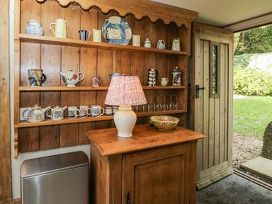 A wooden cabinet with pottery and a lamp next to an open wooden door showing a garden at The Barn at Westhall Cottage Fulbrook near Burford
