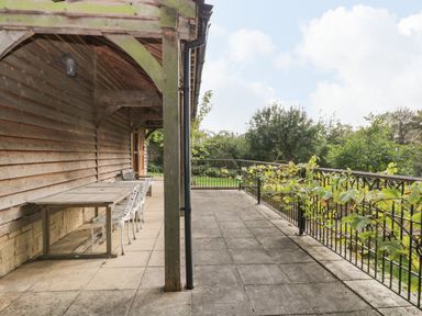 An outdoor covered patio with a wooden table and white metal chairs next to a building with wooden siding at The Barn at Westhall Cottage Fulbrook near Burford