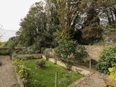 A garden with gravel paths grass a stone planter and a stone wall with trees behind at The Barn at Westhall Cottage Fulbrook near Burford