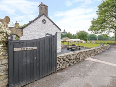A black wooden gate with the sign The Druids stone wall and white building with chimney near a garden with seating and trees at The Druids in Druid near Corwen