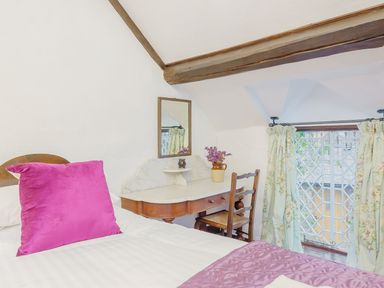 A bedroom with a bed featuring a purple pillow and quilt next to a wooden desk with a mirror and a chair by a window with floral curtains at The Druids in Druid near Corwen