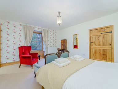 A bedroom with a bed and towels on it a red armchair a wooden door floral wallpaper and a window with curtains at The Druids in Druid near Corwen