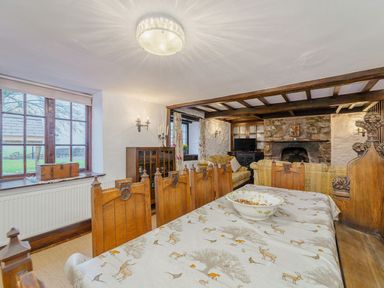 A dining room with wooden chairs and a table covered with a patterned cloth next to a living room with sofas and a stone fireplace at The Druids in Druid near Corwen