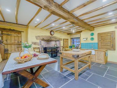 A kitchen with wooden beams on the ceiling a wooden table with bread and fruit a wooden island and a brick fireplace at The Druids in Druid near Corwen