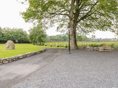 An outdoor area with a gravel seating area under a large tree and a curved paved path beside a grass lawn with a large rock at The Druids in Druid near Corwen