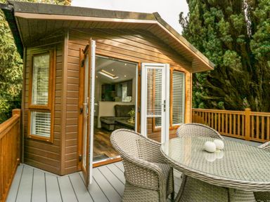A wooden deck with a round glass table and wicker chairs outside a wooden cabin with open doors and large windows