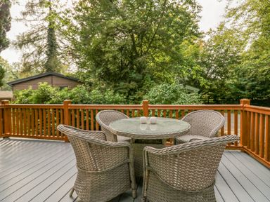 An outdoor deck with four wicker chairs around a round glass table surrounded by trees and wooden railing at Willows Lodge White Cross Bay near Windermere