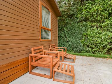Two wooden chairs with footrests and a small table on a tiled patio next to a brown wooden building with a window surrounded by green bushes