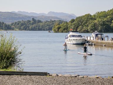 Two people paddleboarding on a lake near boats and a dock with trees and hills in the background at Willows Lodge in White Cross Bay near Windermere