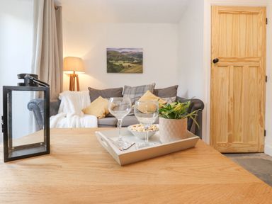 A living room with a wooden table holding a tray with two empty wine glasses a bowl of popcorn and a potted plant a gray sofa with cushions and a white throw a floor lamp and a wooden door at The Wonky Burrow in Bowness-On-Windermere