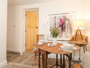 A dining area with a wooden table set with dishes flowers on the table wooden chairs a lamp on a side table and a large framed artwork on the wall at The Wonky Burrow in Bowness-On-Windermere