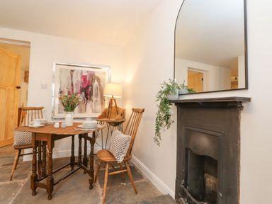 A dining area with a wooden table set with dishes and flowers two wooden chairs with cushions a fireplace with a mirror and greenery a lamp and abstract art on the wall at The Wonky Burrow in Bowness-On-Windermere