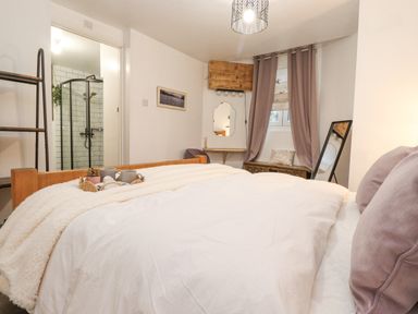 A bedroom with a white bedspread and purple pillows next to a window with purple curtains and a mirror at The Wonky Burrow in Bowness-On-Windermere