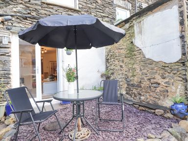 An outdoor patio area with a round glass table under a black umbrella and two black chairs by a stone wall at The Wonky Burrow in Bowness-On-Windermere