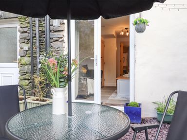 A patio area with a glass table, two black chairs, an umbrella, and flowers in a vase at The Wonky Burrow in Bowness-On-Windermere