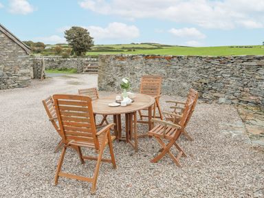 An outdoor seating area with a round wooden table and six wooden chairs on gravel surrounded by stone walls at Caerdegog Ganol in Cemlyn near Cemaes Bay