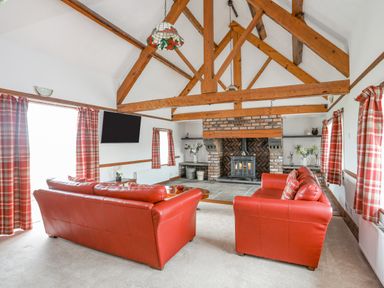 A living room with red leather sofas, a brick fireplace, exposed wooden beams and red plaid curtains at Caerdegog Ganol in Cemlyn near Cemaes Bay
