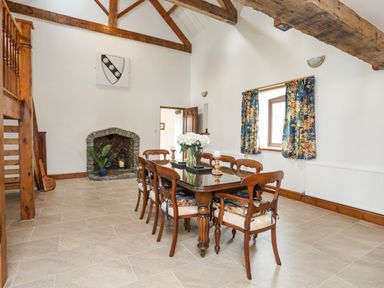 A dining room with a wooden table and chairs near a stone fireplace and floral curtains at Caerdegog Ganol in Cemlyn near Cemaes Bay