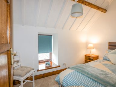 A bedroom with a bed a bedside table with a lamp a window with a blue blind and a chair with a cushion at Caerdegog Ganol in Cemlyn near Cemaes Bay