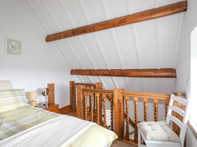 A bedroom with a bed side table lamp and mirror next to wooden railings and a white chair with cushion at Caerdegog Ganol in Cemlyn near Cemaes Bay