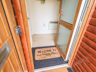 An open wooden front door revealing a welcome mat with text and an interior hallway with a heater on the wall at Our Happy Place Lakeside 16 Borwick near Carnforth
