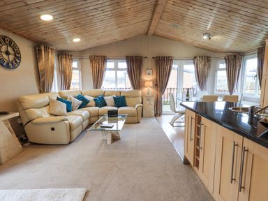 A living room with a beige sectional sofa and blue cushions next to a glass coffee table a dining table with chairs and a kitchen island with a sink at Our Happy Place Lakeside 16 Borwick near Carnforth