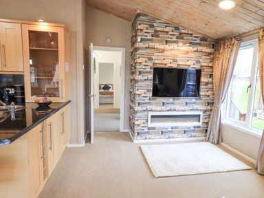 A living area with a TV mounted on a stone wall above a fireplace next to a window with curtains and a kitchen area with cabinets and a countertop at Our Happy Place in Lakeside 16 Borwick near Carnforth