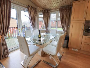 A dining area with a glass table and six white chairs beside windows and wooden cabinets at Our Happy Place Lakeside 16 Borwick near Carnforth