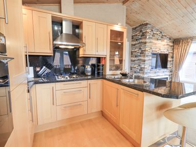 A kitchen with wooden cabinets a black countertop a gas stove a double sink and a sitting area with a stool at Our Happy Place Lakeside 16 Borwick near Carnforth
