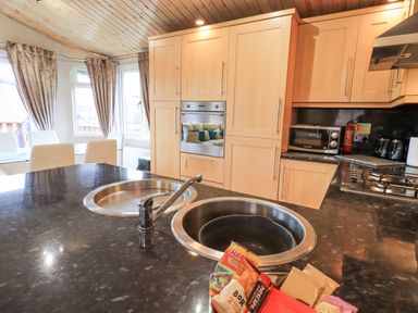 A kitchen with a black countertop island containing two sinks and snacks with wooden cabinets a built in oven a microwave a gas stove and a dining area with white chairs and curtains at Our Happy Place Lakeside 16 Borwick near Carnforth