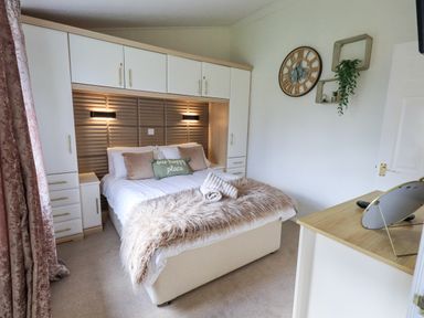 A bedroom with a double bed surrounded by white wardrobes and shelves with a clock and plants on the wall at Our Happy Place Lakeside 16 Borwick near Carnforth