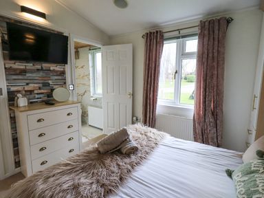 A bedroom with a bed, a dresser with decorative items, a mounted TV, a window with curtains, and a door leading to a bathroom at Our Happy Place in Lakeside 16, Borwick near Carnforth