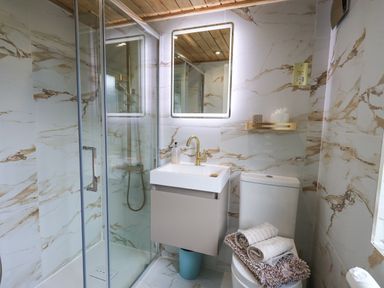 A bathroom with a glass shower enclosure a wall-mounted sink with a faucet a mirror a toilet with towels on top and a shelf with items above the toilet at Our Happy Place Lakeside 16 Borwick near Carnforth