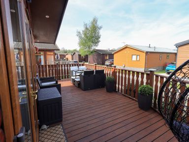 A wooden deck with outdoor seating and planters overlooking wooden cabins and parked cars at Our Happy Place Lakeside 16 Borwick near Carnforth