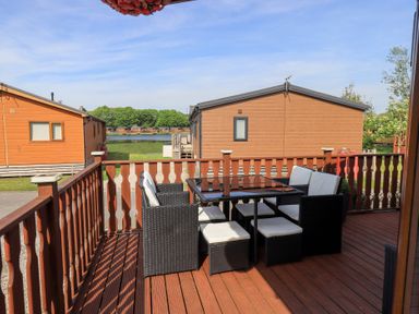 A wooden deck with black wicker chairs and a glass table overlooking brown cabins and a lake at Our Happy Place Lakeside 16 Borwick near Carnforth