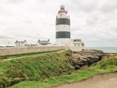 A lighthouse with black and white horizontal stripes near the sea with green grass and rocky cliffs at Old Field House Slade near Fethard-On-Sea County Wexford