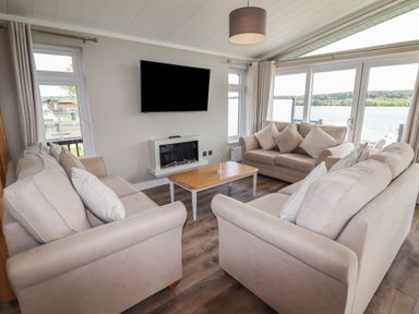 A living room with three beige sofas, a wooden coffee table, a wall-mounted TV, and a fireplace with large windows overlooking a lake at 40 Delamere Point in Delamere